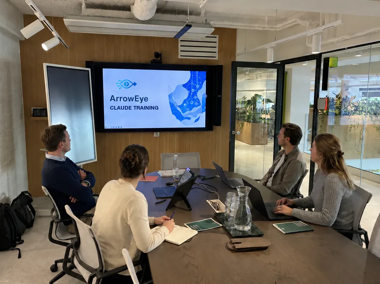 Four participants at a meeting table look at the screen showing the opening slide of the ArrowEye Claude training
